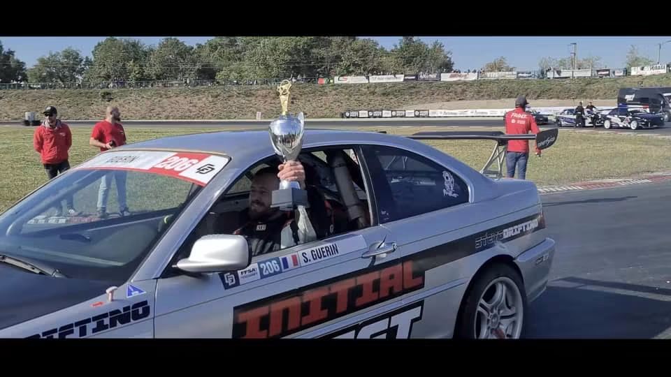 BMW Initial DRIFT argentée et noire ; un trophée est tenu par la fenêtre côté pilote.